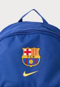 Blå ryggsäck med FC Barcelonas emblem och gul Nike swoosh-logotyp på framfickan, övre handtag synligt mot vit bakgrund.