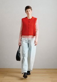 Crveni prsluk sa rebrastim uzorkom i gumbima preko prugaste košulje dugih rukava, u paru sa svijetloplavim hlačama i crnim cipelama, drži crnu torbu.