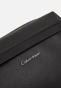 Černá texturovaná kožená psaníčko s hladkým černým páskem a stříbrným logem "Calvin Klein" na přední straně. Obdélníkový tvar.