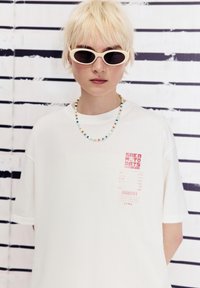 Witte oversized t-shirt met een gedurfde rode grafische label. Draagt met een kleurrijke kralen ketting en witte cat-eye zonnebril. Achtergrond met verticale strepen.