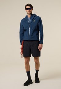 Hombre con gafas de sol, chaqueta azul con cremallera, pantalones cortos negros, calcetines y zapatos negros, sosteniendo una cuerda de escalada roja.