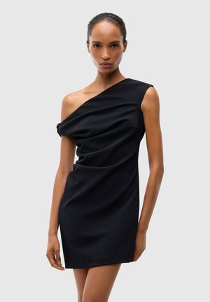 Donna che indossa un vestito corto nero monospalla con tessuto drappeggiato, in piedi davanti a uno sfondo chiaro e semplice.