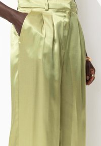 Pantalons en satin vert clair à taille haute, avec des poches latérales et des détails plissés à l'avant, mettant en valeur une texture lisse et brillante.