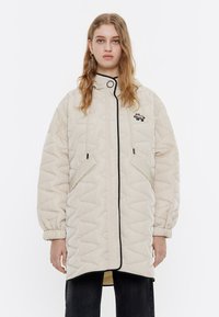 Chaqueta acolchada beige con ribete negro, cuello alto y bolsillos laterales. Presenta un logo en el pecho y puños elásticos.