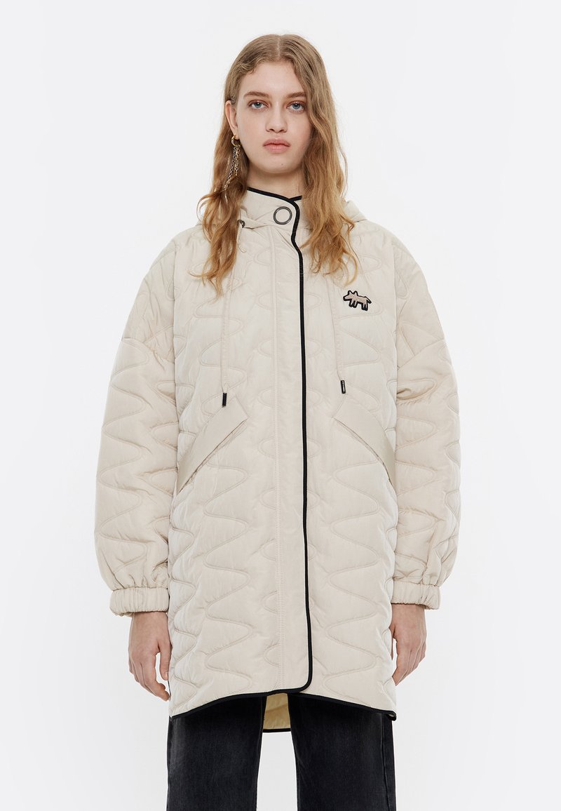 Chaqueta acolchada beige con ribete negro, cuello alto y bolsillos laterales. Presenta un logo en el pecho y puños elásticos.