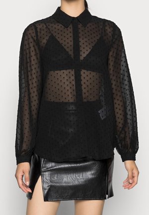 Vrouw draagt een doorschijnende zwarte blouse met witte stippen over een zwarte bralette en een zwarte leren minirok met een splitsing aan de voorkant, staande tegen een neutrale achtergrond.