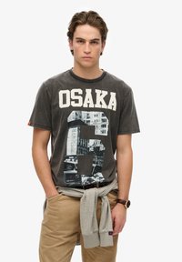 Camiseta de algodón gris con gran texto "OSAKA" en blanco, impresión gráfica de un paisaje urbano, cuello redondo y mangas cortas, combinada con pantalones caqui.