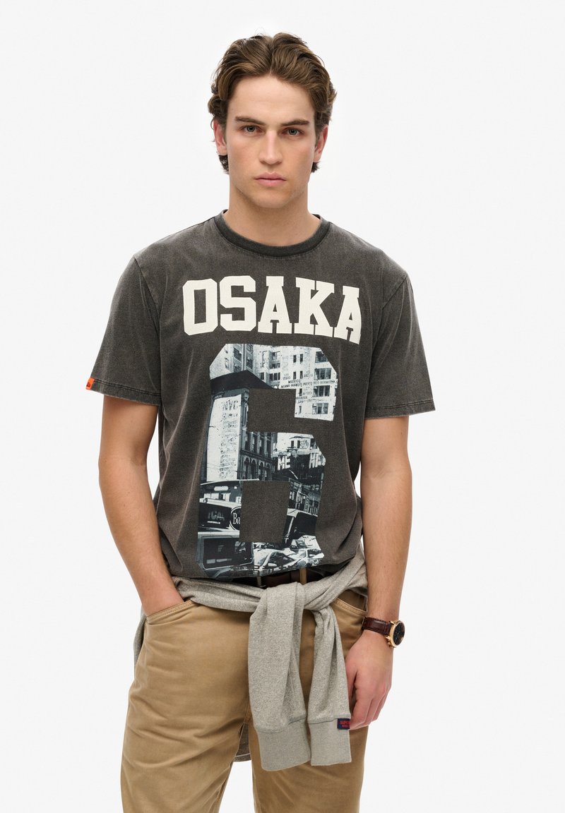 Camiseta de algodón gris con gran texto "OSAKA" en blanco, impresión gráfica de un paisaje urbano, cuello redondo y mangas cortas, combinada con pantalones caqui.