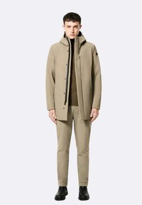 Beige waterdichte jas met capuchon, volledige rits en drukknopen. Gecombineerd met bijpassende beige broek en zwarte enkellaarsjes. Gladde textuur.