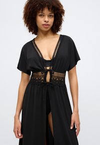 Μαύρο κάλυμμα με κοντά μανίκια, δαντελένια διακόσμηση στη μέση και κλείσιμο με κορδόνι. Ελαφρύ ύφασμα με ροή.