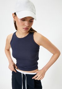 Top sin mangas de color marino con un escote redondo, combinado con pantalones oscuros que tienen un cordón blanco. El modelo lleva una gorra de color gris claro.