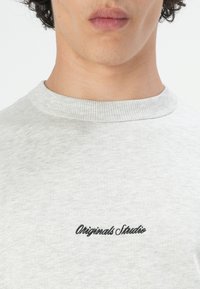 Lichtgrijze sweatshirt met een ribbelkraag, met zwarte geborduurde tekst "Originals Studio" op de borst. Zachte textuur.