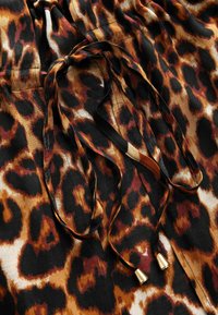 Tkanina s leopard uzorom u nijansama smeđe, crne i kreme; sadrži vezicu s zlatnim metalnim vrhovima i mekani je tekstura.