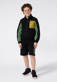 Petit garçon aux cheveux bouclés portant une veste noire avec des panneaux verts et jaunes, un short noir et des baskets noires, se tenant sur un fond blanc.