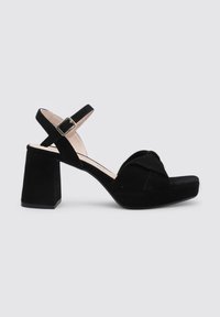 Sandalias de tacón bloque en ante negro con una correa ancha, hebilla en el tobillo y diseño frontal anudado. La suela gruesa añade altura y estabilidad.