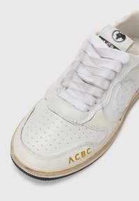 Zapatilla de cuero blanca con puntera perforada, superficie lisa, cordones grises y logo dorado "ACBC" en el lateral. Detalles de la suela de goma visibles.