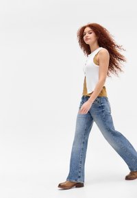 Hvit ermeløs topp lagt over en sennepsfarget tank, parret med lyseblå flagrende jeans og brune ankelboots. Langt krøllete hår som faller nedover.