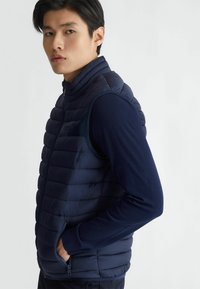 Gilet puffer navy con design a righe orizzontali e colletto alto, dotato di zip frontale e tasche laterali, indossato sopra una maglietta a maniche lunghe.