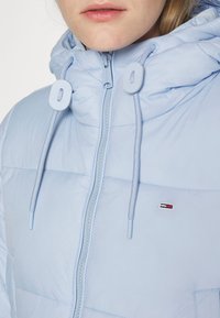 Detailní záběr osoby oblečené v světle modré vycpané bundě se zipem a kapucí, která má nastavitelné šňůrky a malé obdélníkové logo na hrudi.