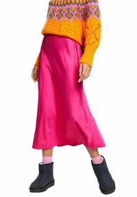Jupe midi en satin rose à la texture lisse, associée à un pull orange vif et des bottines noires. La tenue combine des couleurs audacieuses.