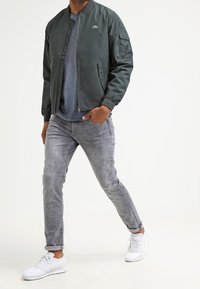 Chaqueta bomber verde oscuro con cierre frontal de cremallera, bolsillos laterales y bolsillo en el brazo. Lleva puesta sobre una camiseta azul, combinada con jeans gris claro y zapatillas blancas.