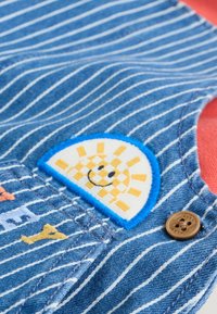 Blåstribet denimstof med et flerfarvet broderet applikation, der viser en smilende sol på en hvid baggrund samt en brun knapdetalje.