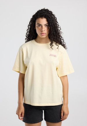 Jonge vrouw met lang krullend haar die een lichtgeel T-shirt draagt bedrukt met "Hawaii" en zwarte korte broek, staand tegen een effen achtergrond.