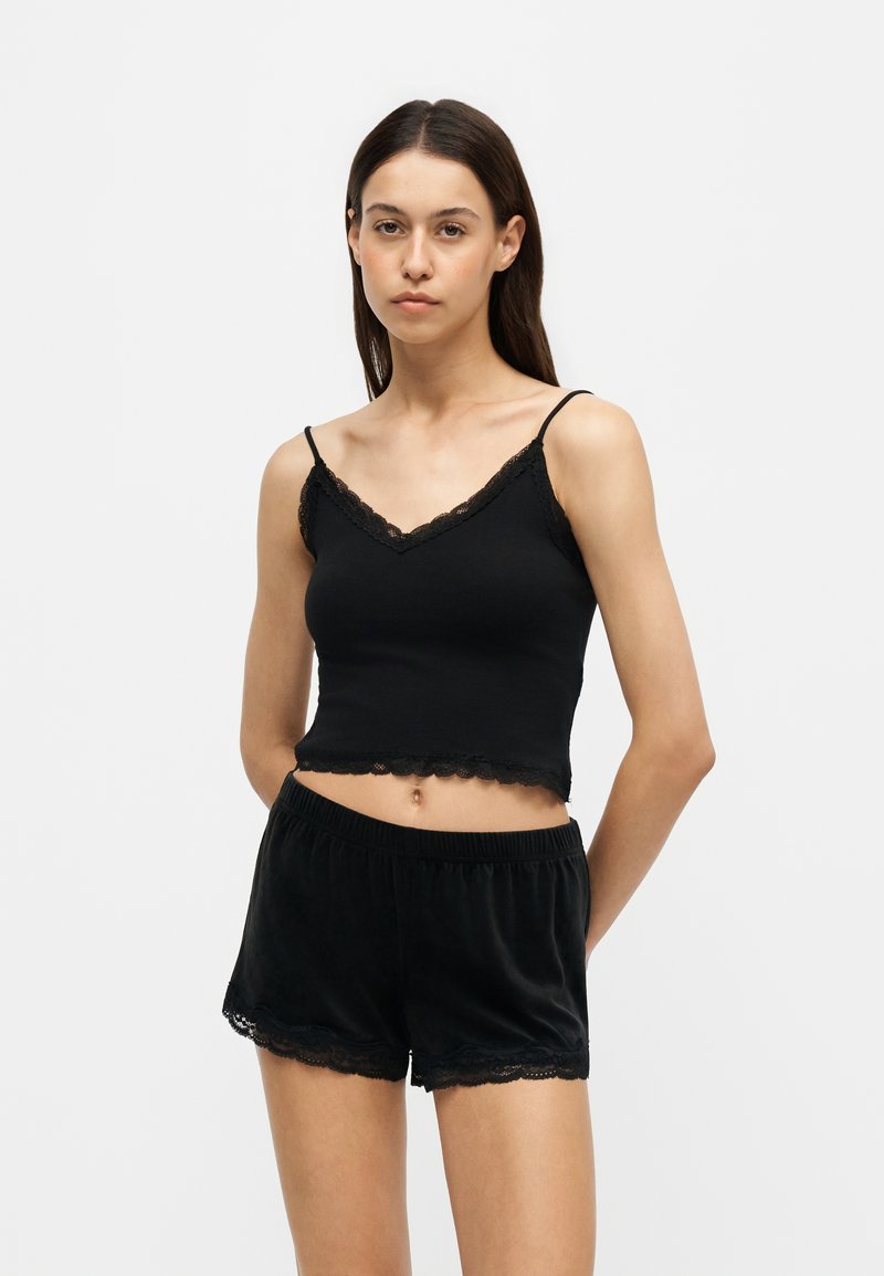 Jeune femme aux longs cheveux foncés portant un crop top noir sans manches orné de dentelle et un short assorti orné de dentelle sur fond blanc.