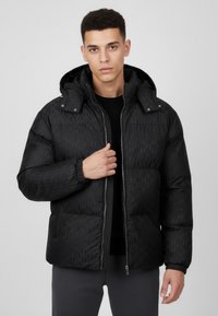 Svart vadderad jacka med huva, med texturerad yta och diagonalt mönster, dragkedja framtill och elastiska muddar, bärs över en svart tröja.