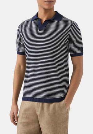 Uomo che indossa una polo a maniche corte a righe blu navy e bianco con colletto blu navy e pantaloni beige, una mano in tasca, su sfondo uniforme.