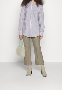 Donna che indossa una camicia a maniche lunghe a righe verticali, pantaloni beige cropped, stivali a calza e tiene una piccola borsa fluffy verde menta.