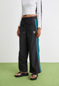 Black track pants with a turquoise side stripe, elastic waistband, and Puma logo; paired with black flat sandals on green surface.