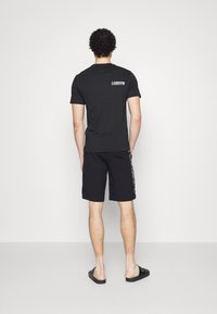 Homme vu de dos portant un t-shirt noir avec un petit logo blanc, un short noir avec du texte sur le côté et des sandales noires.