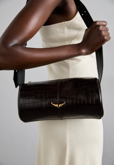 Hnedá kožená taška s krokodílím reliéfom, valcovitého tvaru, s pozláteným detailom a dlhým čiernym popruhom, ktorú drží osoba.