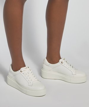 Personne portant des baskets compensées blanches avec des lacets, debout sur une surface gris clair.