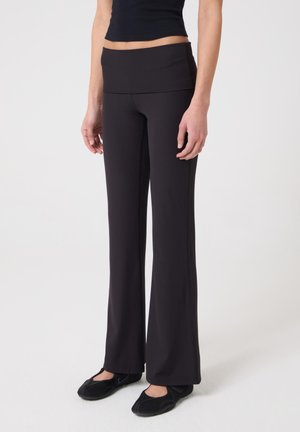 Mujer con pantalones negros de talle alto y piernas anchas y zapatos negros sin cordones, de pie contra un fondo claro y liso.