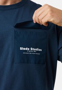 T-paita tummansinisestä kankaasta, jossa on neliön muotoinen tasku, jossa on teksti. Materiaali on pehmeä ja siinä on matta pinta. Taskussa on kontrastikasvi.