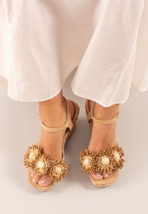 Sandalias de cuña en tono beige con pompones decorativos en color crema y detalles de madera. Tienen una correa alrededor del tobillo y una plantilla acolchada.