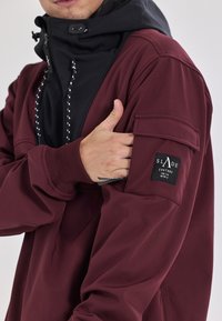 Bordeaux jakke med sort hætte, med lynlås foran, justerbare snore og et logo patch på venstre ærme. Glat tekstur.