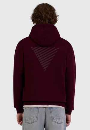 Person wearing a dark purple hoodie with a large striped triangle design on the back, paired with light blue jeans.