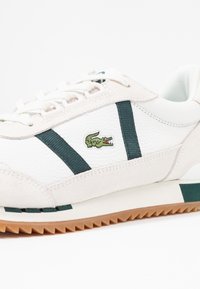 Baskets en cuir blanc et daim avec des rayures vertes et un logo crocodile brodé en vert. Semelle en caoutchouc texturée avec des bords dentelés.