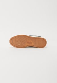 Black sneaker sole with textured rubber, light brown accents, and embossed logo, featuring ridges for grip and a circular pattern.
