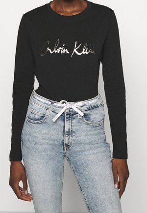 Osoba nosząca czarną koszulkę z długim rękawem z logo „Calvin Klein” oraz jasnoniebieskie jeansy z wysokim stanem i białym firmowym paskiem ze sznurkiem.