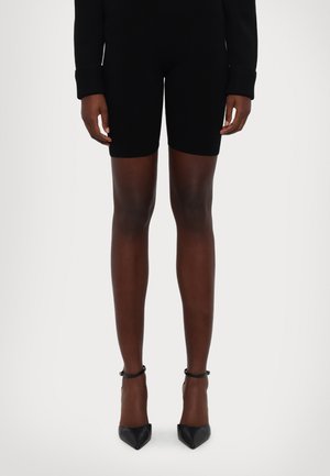 Body in maglia nera a maniche lunghe, abbinato a pantaloncini neri aderenti. Il modello indossa tacchi neri a punta con cinturini alla caviglia.
