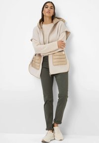 Beige Kapuzenpullover mit strukturiertem Strick und gepolsterten, tanfarbenen Fronttaschen, kombiniert mit olivgrünen Hosen und hellbeigen Sneakers.