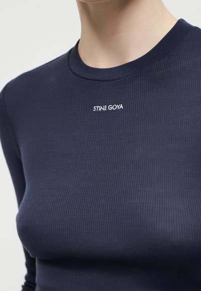Κοντινό πλάνο ενός ατόμου που φοράει μπλε σκούρο ριμπ μακρυμάνικη μπλούζα με κεντημένο το "STINE GOYA" σε λευκό στο κέντρο του στήθους.