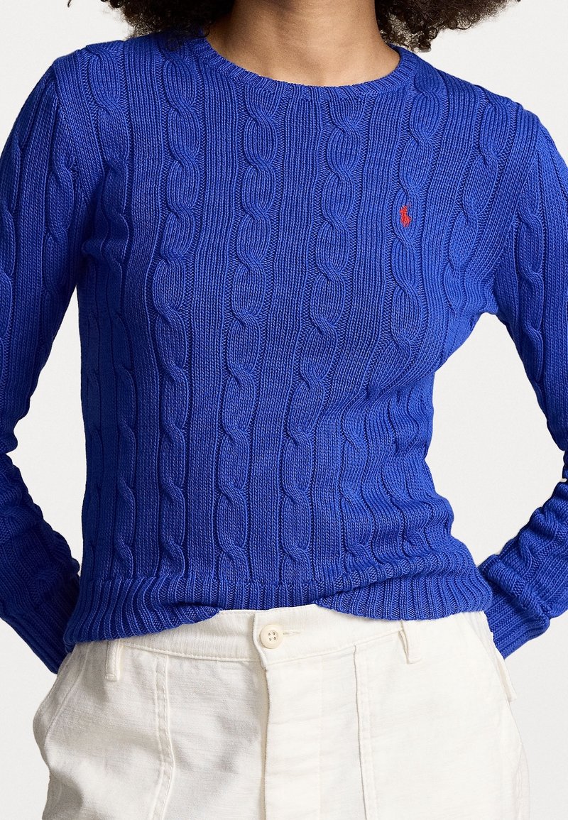 Personne portant un pull torsadé bleu vif avec un petit logo rouge, associé à un pantalon taille haute blanc cassé.