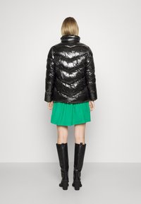 Chaqueta de plumas negra y brillante con costuras en forma de chevrón, cuello alto, combinada con una falda verde y botas negras hasta la rodilla.