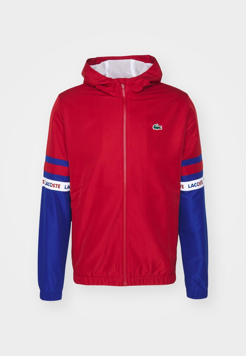 Rood-blauwe windbreakerjas met een capuchon, volledige rits, elastische manchetten en "LACOSTE" merknaam op de mouwen. Bevat een klein logo op de borst.