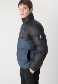Polstret jakke med høy krage, farget i sort og marineblå med et Calvin Klein-logo merke på ermet, og side-lommer.
